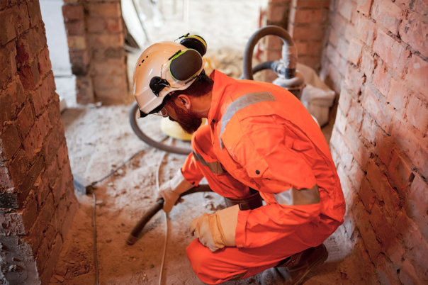 Männliche Reinigungskraft in orangenem Overall und mit Bauhelm reinigt auf Baustelle mit Schlauch